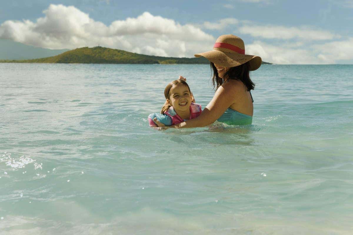Una madre con sombrero sujetando a una niña nadando en una playa de aguas cristalinas