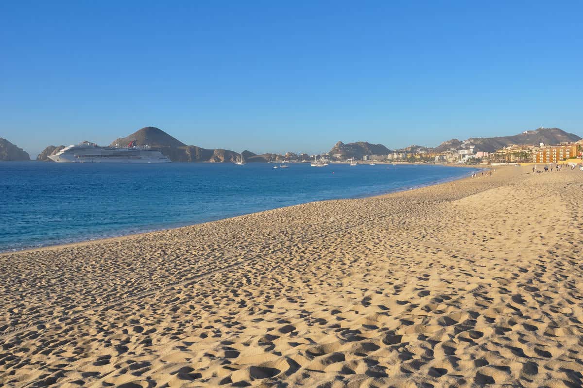Una amplia y dorada playa de arena que se extiende hacia una bahía azul donde un gran barco de crucero está anclado. Colinas bajas y el pueblo costero son visibles en la distancia. El cielo es de un azul claro y uniforme