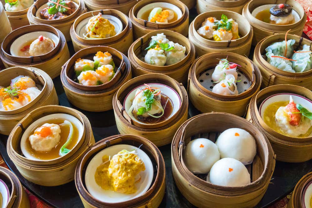 Vista dall'alto di una ricca tavolata ricoperta di cestelli di bambù per la cottura a vapore, ciascuno contenente diverse varietà di dim sum cinesi, tra cui baozi morbidi, siu mai e ravioli trasparenti con guarnizioni colorate