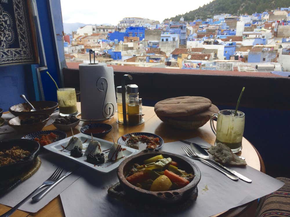 Mesa preparada para comer en una terraza con vistas en Chefchaouen, con platos tradicionales marroquíes entre ellos un tajín de verduras, queso de cabra, pan árabe y bebidas, con la característica ciudad azul y sus colinas al fondo