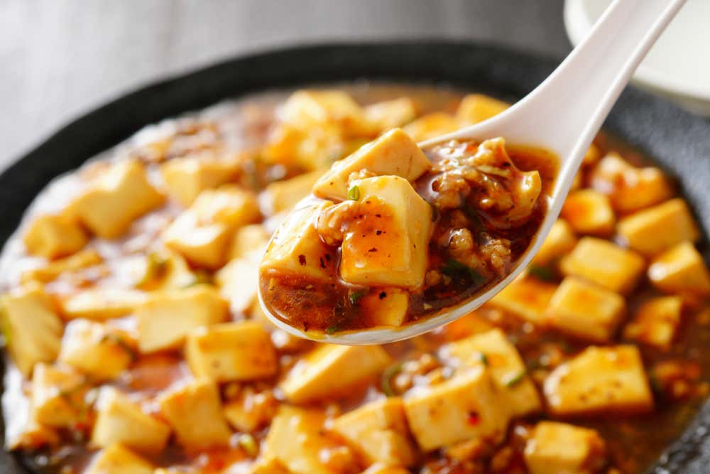 Primo piano di un cucchiaio da zuppa in ceramica bianca che raccoglie una porzione di Mapo Tofu, piatto cinese a base di cubetti di tofu morbidissimo e carne macinata immersi in una salsa rossa e piccante al pepe del Sichuan