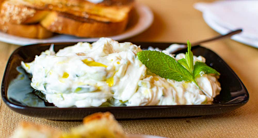 Un primo piano di un piatto di tzatziki greco autentico, servito in una ciotola nera su un tavolo da taverna. La salsa a base di yogurt cremoso, cetrioli e aglio è condita con olio d'oliva e una guarnizione di menta fresca. Sullo sfondo si vede un piatto di pane pita tostato