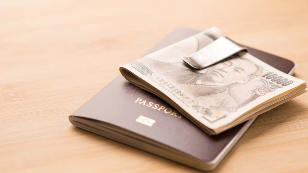 Un passaporto marrone scuro appoggiato su una superficie in legno chiaro, su cui è appoggiata una mazzetta di banconote da 10.000 yen giapponesi piegate e tenute insieme da un fermasoldi in metallo argentato