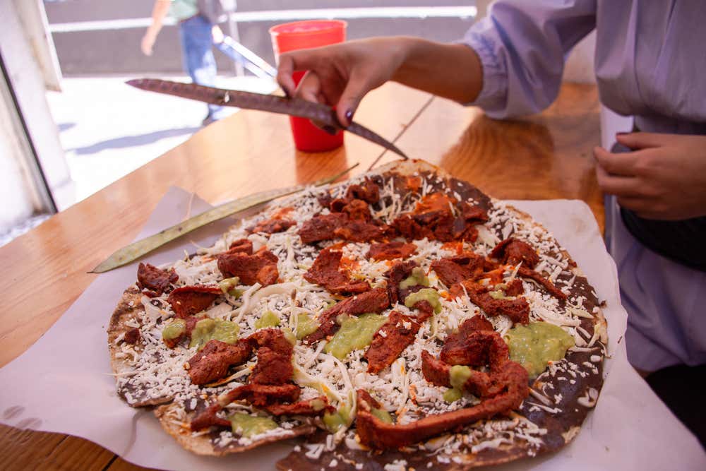 A shot of a Tlayuda being prepared with various toppigns and cheese in a restaurant.