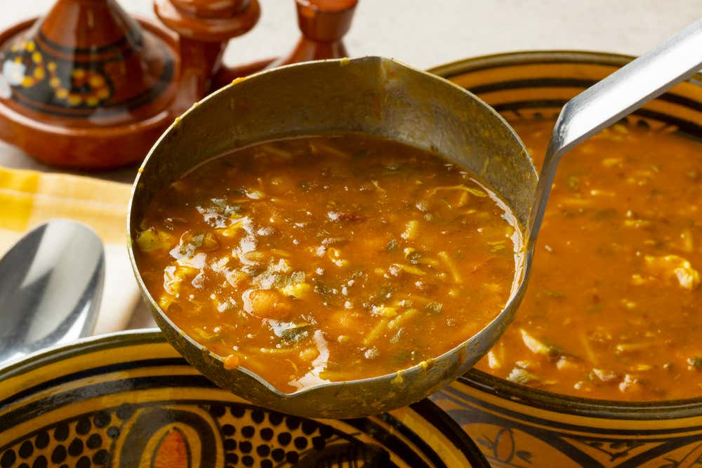 Un cucharón de metal lleno de harira, la densa sopa tradicional marroquí a base de tomate, lentejas, garbanzos y hierbas aromáticas, levantado sobre un cuenco de cerámica finamente decorado, con un tajín de barro al fondo