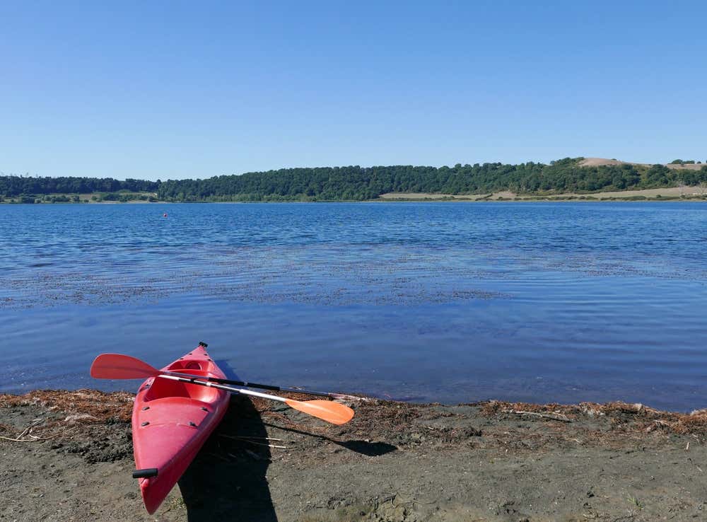 Un kayak rosso con una pagaia adagiata sopra posizionato sulla riva del Lago di Martignano, con le calme acque blu del lago e le colline verdi della riserva naturale sullo sfondo sotto un cielo sereno