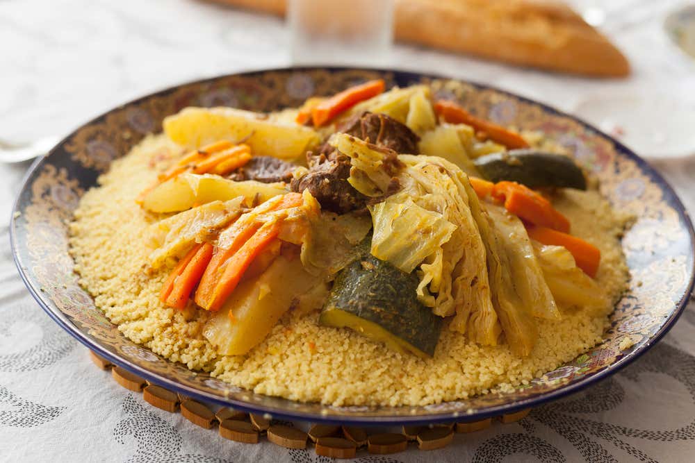 Plato tradicional de cuscús servido en una bandeja de cerámica decorada, con trozos de carne en el centro y verduras cocidas como zanahoria, calabacín, col y patata