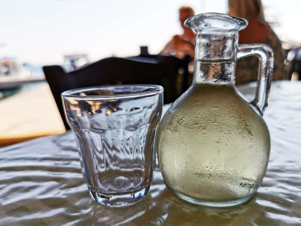 Una caraffa di vetro appannata e ben fredda piena di vino retsina greco, affiancata da un bicchiere vuoto su un tavolo da taverna all'aperto vicino al mare