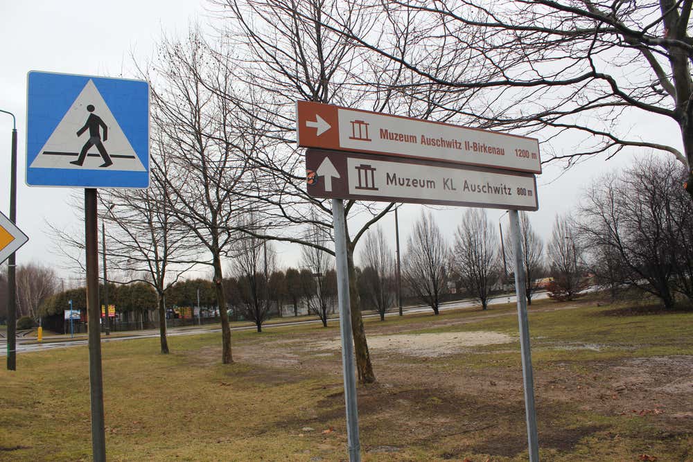 Cartelli stradali turistici marroni che indicano le direzioni e le distanze per il "Muzeum KL Auschwitz" (800 m) e il "Muzeum Auschwitz II-Birkenau" (1200 m), affiancati da un segnale di attraversamento pedonale su uno sfondo alberato autunnale