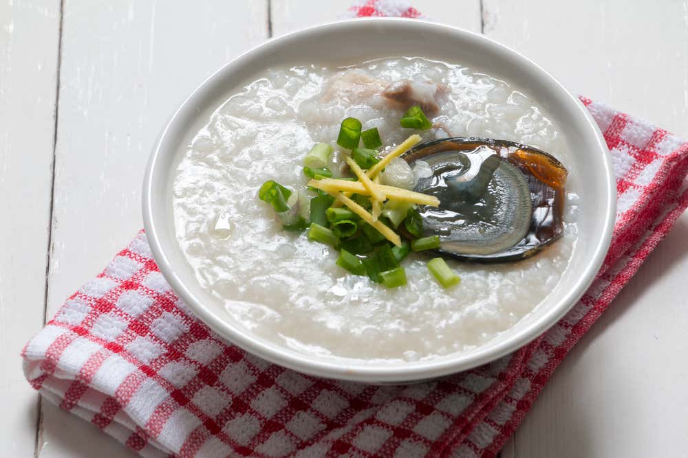 Una ciotola bianca di congee cinese (porridge di riso) servita con maiale, mezza uovo dei cent'anni scura, zenzero a fiammifero ed erba cipollina fresca tritata, adagiata su un canovaccio a scacchi rossi e bianchi