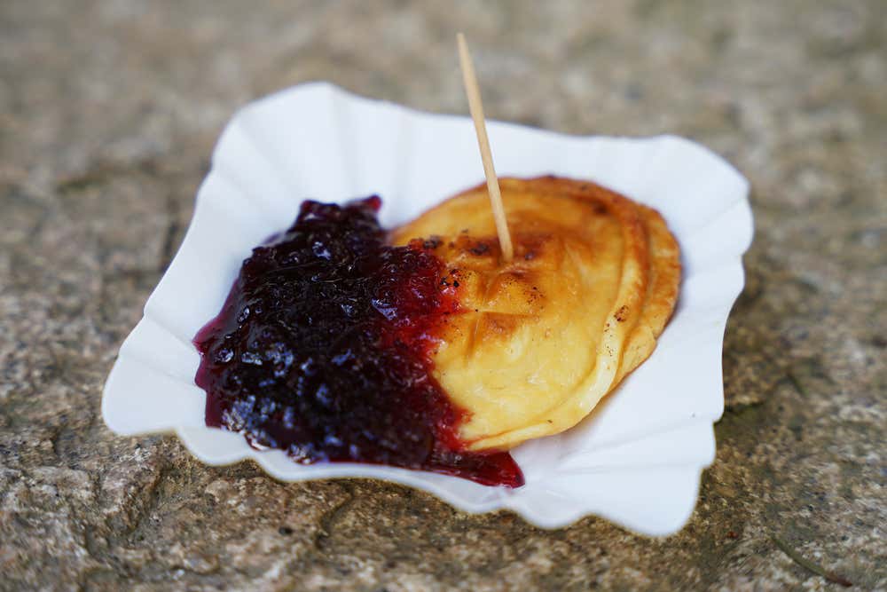 Una porzione di formaggio oscypek grigliato con decorazioni tradizionali incise sulla superficie, servito caldo in un piattino di carta bianco con uno stuzzicadenti e accompagnato da una generosa cucchiaiata di marmellata di mirtilli rossi