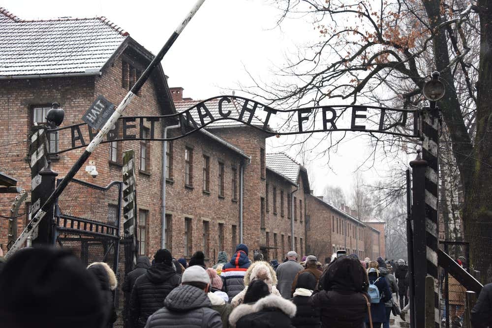 Gruppo di visitatori visti di spalle che attraversano l'iconico cancello d'ingresso del campo di concentramento di Auschwitz I sotto la nota scritta in ferro battuto "Arbeit Macht Frei", circondati da edifici in mattoni rossi e un paesaggio invernale