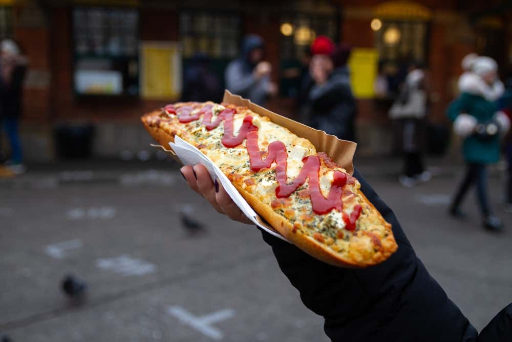 Una mano regge una zapiekanka calda e croccante, la tipica mezza baguette polacca cotta al forno e farcita con formaggio fuso, funghi ed erbe aromatiche, completata da una spirale di ketchup sulla superficie, in un contesto urbano all'aperto