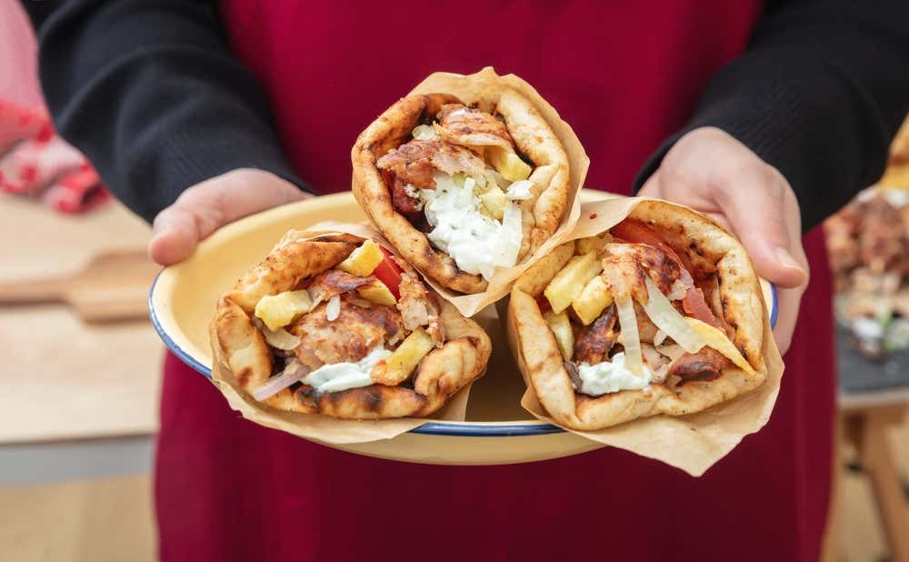 Una persona con grembiule rosso regge un vassoio giallo contenente tre pita gyro tradizionali avvolte nella carta, farcite con carne grigliata, patatine fritte, salsa tzatziki, pomodori e cipolla