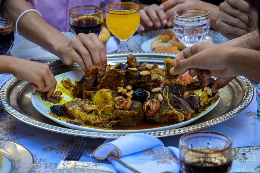 Primo piano di un piatto tradizionale marocchino di carne di agnello stufata con prugne, albicocche secche, mandorle e noci, servito al centro della tavola su un vassoio d'argento decorato e condiviso con le mani da diverse persone