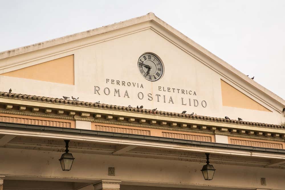 Facciata storica in stile classico dell'ingresso della stazione ferroviaria della Roma-Lido a Piramide, caratterizzata da un frontone triangolare bianco con un orologio analogico al centro e le iscrizioni "FERROVIA ELETTRICA" e "ROMA OSTIA LIDO" in rilievo