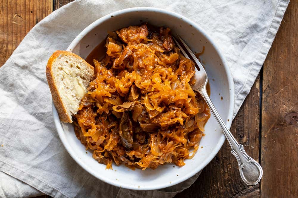 na ciotola bianca vista dall'alto contenente bigos polacco, uno stufato ricco di cavoli, crauti e carne, servito con una fetta di pane spalmata di burro e una forchetta d'argento, adagiato su un tovagliolo chiaro e un tavolo di legno