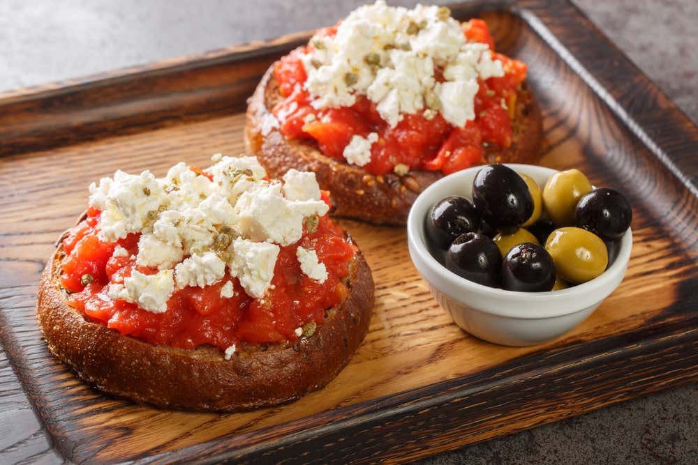 Due ntakos cretesi disposti su un vassoio di legno scuro, preparati con base di frisa d'orzo, pomodori maturi a cubetti, formaggio mizithra sbriciolato e origano, affiancati da una ciotolina di olive nere e verdi