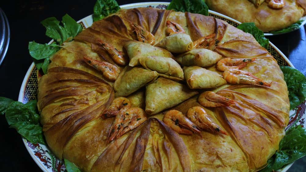 Una pastela de pescado tradicional marroquí servida en un plato decorado, hecha con hojaldre dorado y crujiente dispuesto en pétalos, decorada por encima con gambas enteras y fardos rellenos (briouates) triangulares, con hojas de lechuga en los bordes