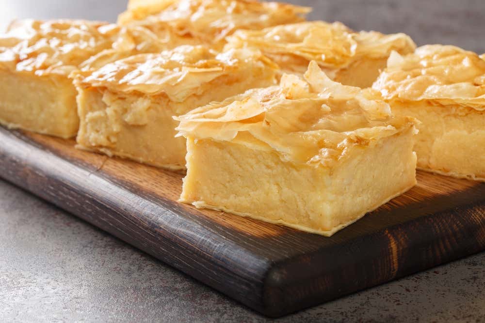 Quadrotti di galaktoboureko disposti su un vassoio di legno scuro, dolce greco a base di uno spesso strato di crema di semolino racchiuso tra fogli di pasta fillo dorata e fragrante, impregnata di sciroppo