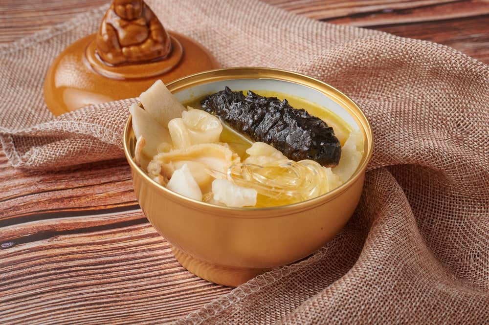 Una ciotola dorata contenente la zuppa prelibata "Buddha salta il muro", con ingredienti pregiati come cetriolo di mare scuro, orecchia di mare e pinna di squalo, accostata al suo coperchio sormontato da una statuetta di Buddha