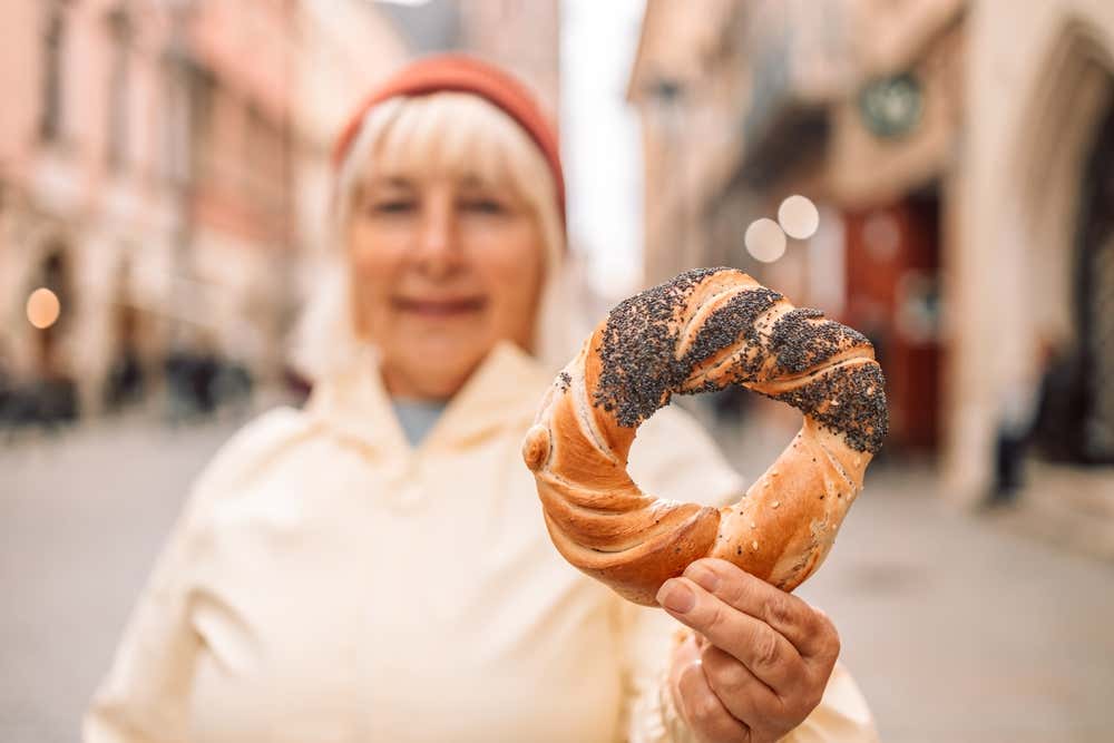 Una donna in vestiti invernali tiene in mano, e mostra in primo piano, una pagnotta intrecciata di forma circolare decorata con semi di papaveri, uno snack tipico di Cracovia, Sullo sfondo i palazzi del centro storico di Cracovia in una giornata autunnale