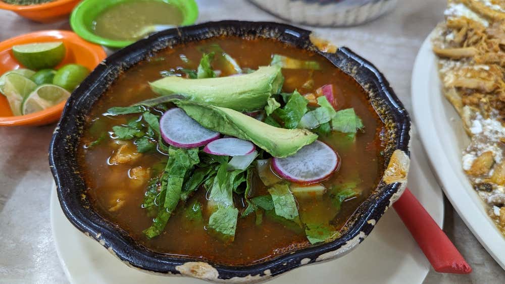 A bowl containing Pozole topped with horse radish, avocado and lettuce.