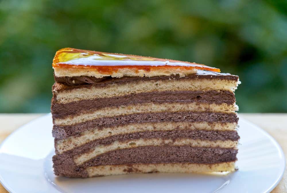 Primo piano di una fetta di torta Dobos su un piattino bianco, con i suoi caratteristici strati alternati di pan di Spagna e crema al cioccolato, sormontata da un sottile e lucido strato di caramello dorato