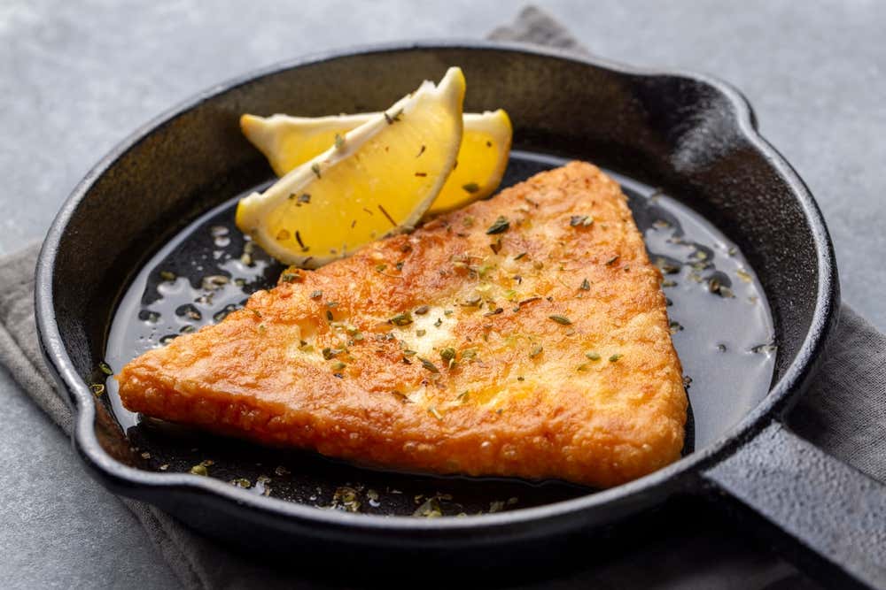 Formaggio saganaki fritto fino a doratura, di forma triangolare e spolverato di origano, servito caldo in un padellino di ghisa nero con due spicchi di limone fresco