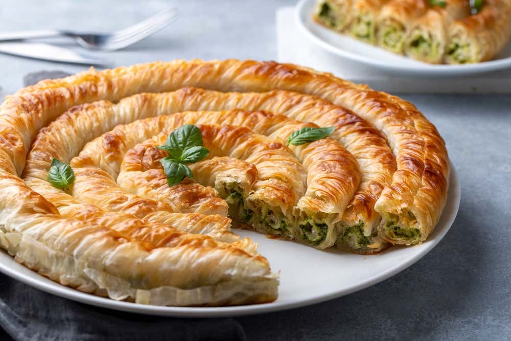 na torta salata tradizionale greca (spanakopita) a forma di spirale dorata e croccante, fatta di pasta fillo arrotolata e farcita con spinaci e formaggio feta, servita su un piatto bianco con una fetta tagliata che mostra il ripieno