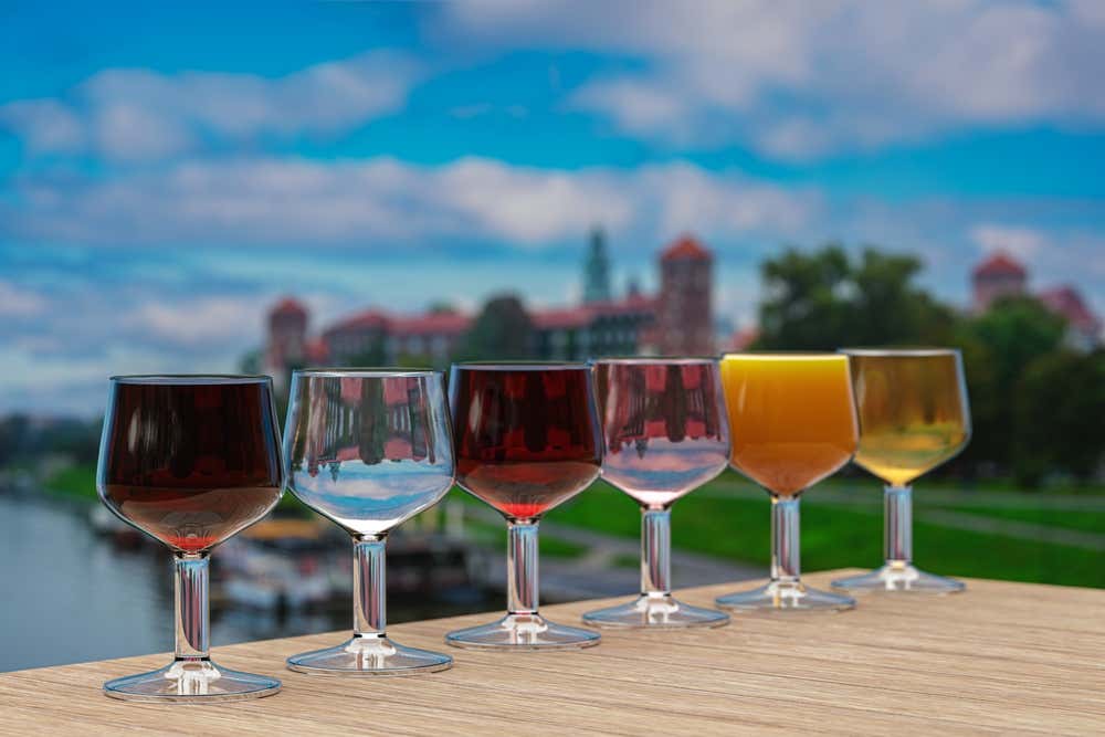 Una fila di sei bicchierini da liquore posizionati su un tavolo di legno all'aperto, riempiti con diversi tipi di nalewka polacca dai colori vari (dal rosso scuro al trasparente, rosa, arancione e dorato), con lo sfondo sfocato del castello del Wawel a Cracovia