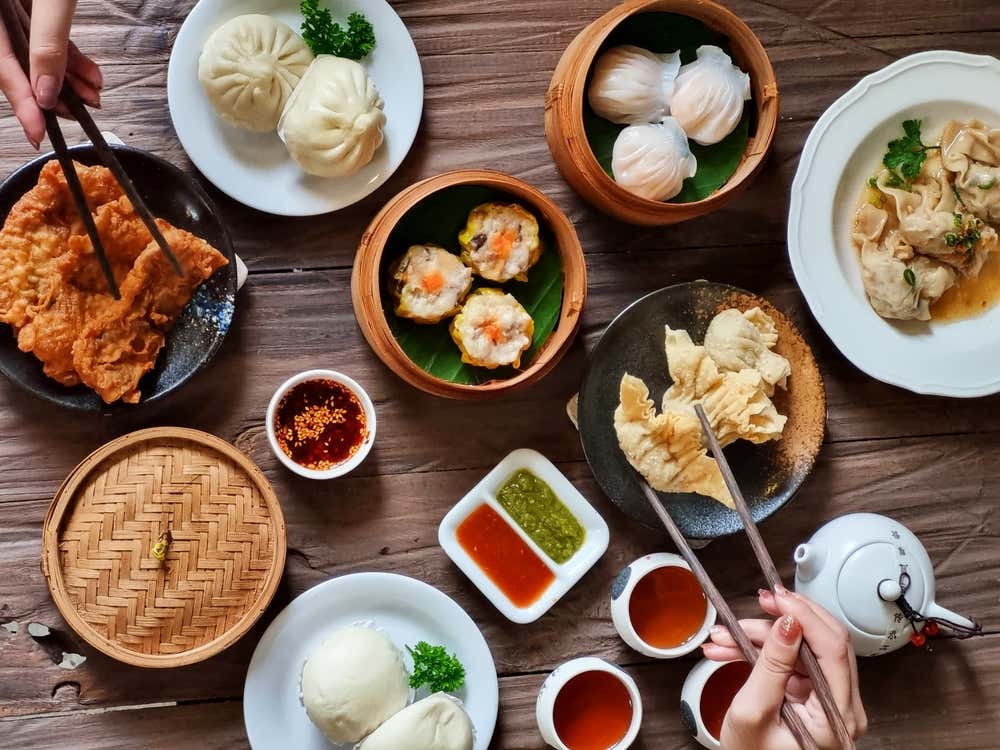 Vista dall'alto di un tavolo di legno imbandito con una selezione di piatti cinesi e dim sum, tra cui baozi al vapore, ravioli har gow, siu mai, ravioli fritti e ciotoline con salse di accompagnamento