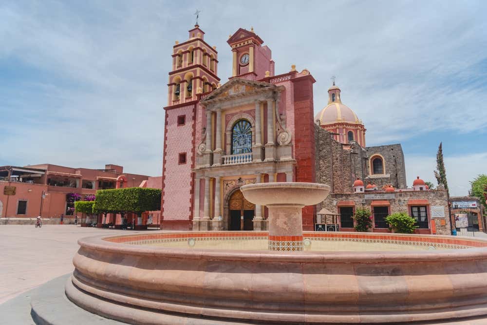 The Church Parroquia Santa Maria de Asuncion with a fountain in the foreground and a cloudy sky in the background.