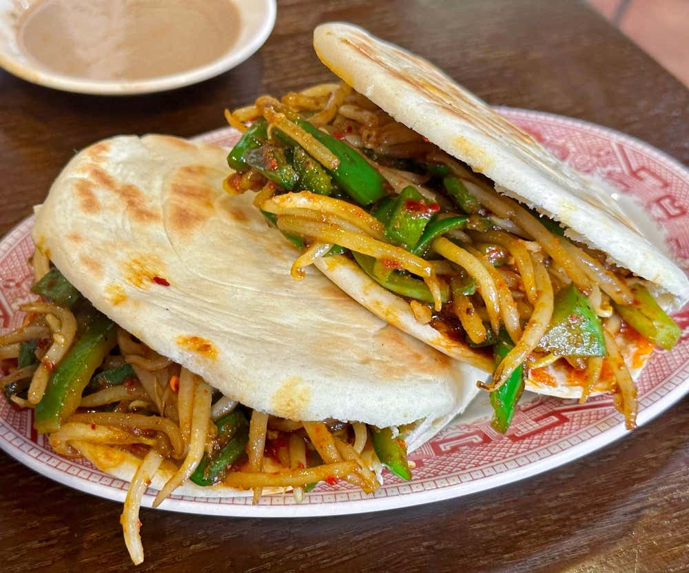 Due panini cinesi piatti e dorati alla piastra (Rou Jia Mo in versione vegetariana), morsi o aperti, ripieni di germogli di soia, peperoni verdi saltati e spezie piccanti su un piatto decorato.