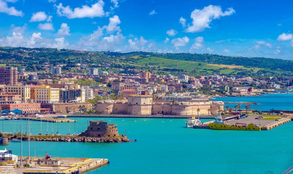 Vista panoramica del porto di Civitavecchia con l'imponente Forte Michelangelo in pietra al centro, il Forte del Lazzaretto su un molo in primo piano, navi e barche ormeggiate nelle acque turchesi e le colline verdi dello sfondo sotto un cielo azzurro con nuvole sparse
