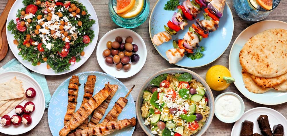 Vista dall'alto di un tavolo di legno imbandito con piatti della cucina mediterranea e greca, tra cui spiedini di pollo grigliati, insalata di cavolo riccio e ceci, insalata di pasta con feta, pane pita, olive e salse
