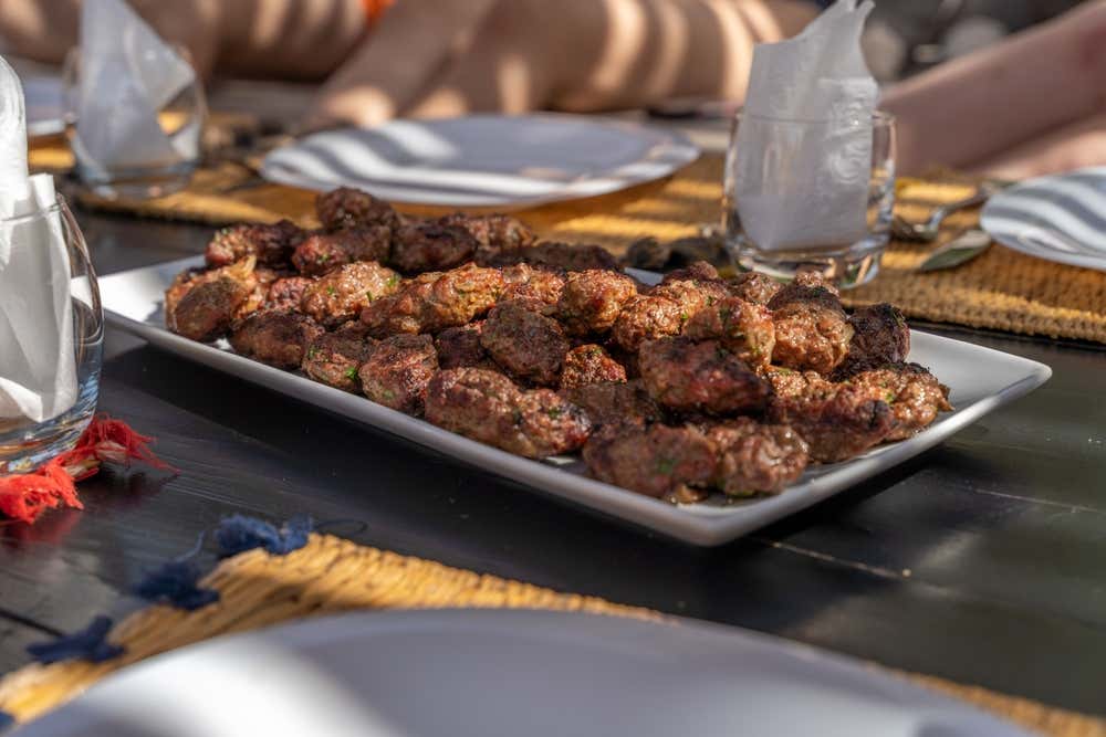 Una bandeja llena de pinchos de carne especiada, colocada sobre una mesa preparada para la cena, con las piernas de algunos comensales al fondo