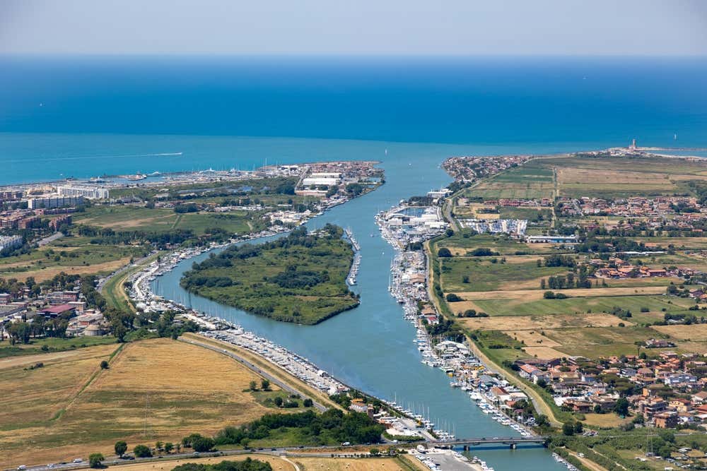 Veduta aerea della foce del fiume Tevere presso Fiumicino e Ostia, con l'Isola Sacra verdeggiante al centro separata dai due bracci d'acqua del fiume, numerosi moli e imbarcazioni ormeggiate lungo le sponde, e il mare Tirreno blu all'orizzonte.