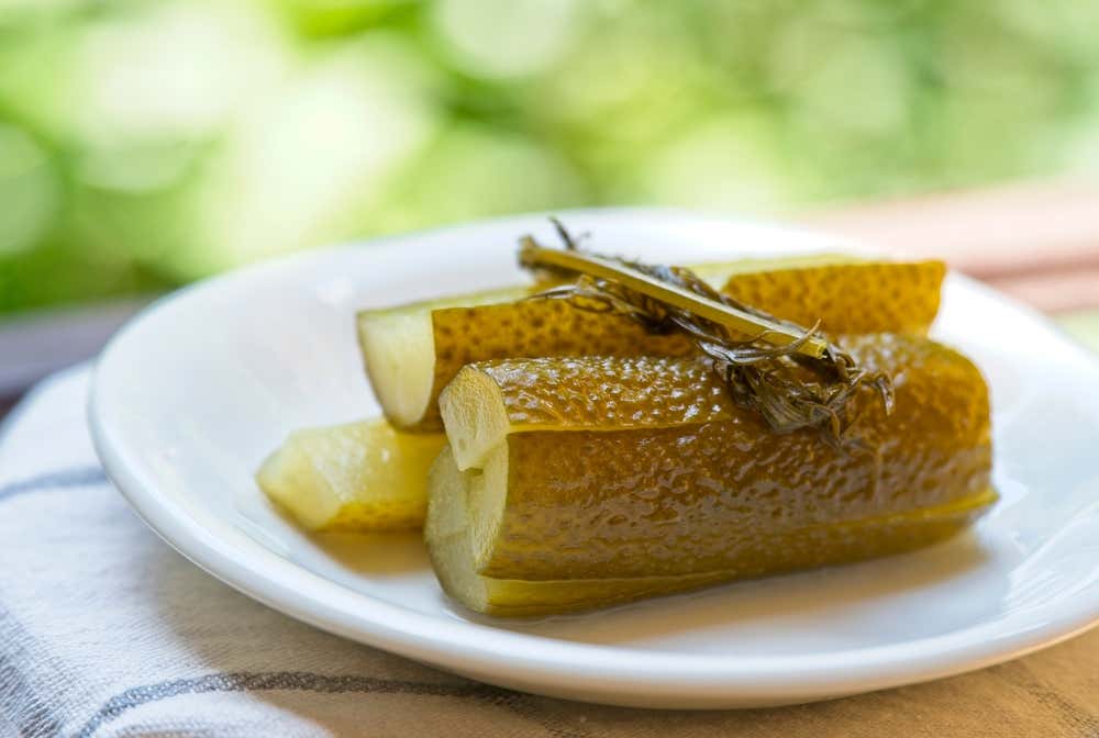 Primo piano di cetriolini sott'aceto tagliati a metà nel senso della lunghezza, serviti su un piattino bianco e guarniti con aneto, con uno sfondo verde sfocato