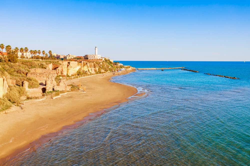 Veduta panoramica della spiaggia della Villa di Nerone ad Anzio, con resti romani in mattoni lungo la falesia dorata, la sabbia chiara che curva verso il mare cristallino sfumato di azzurro e il faro di Capo d'Anzio sullo sfondo sotto un cielo sereno