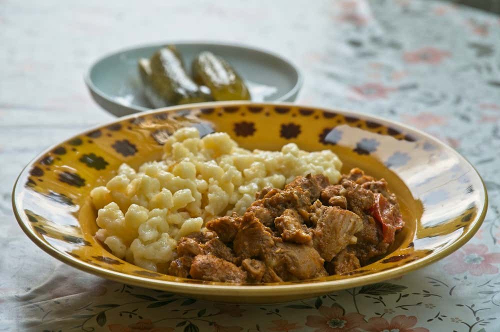 Piatto fondo in ceramica decorata contenente speätzle o nokedli da un lato e uno stufato di carne spezzatino (pörkölt) dall'altro, accompagnato sullo sfondo da un piattino con cetriolini sott'aceto