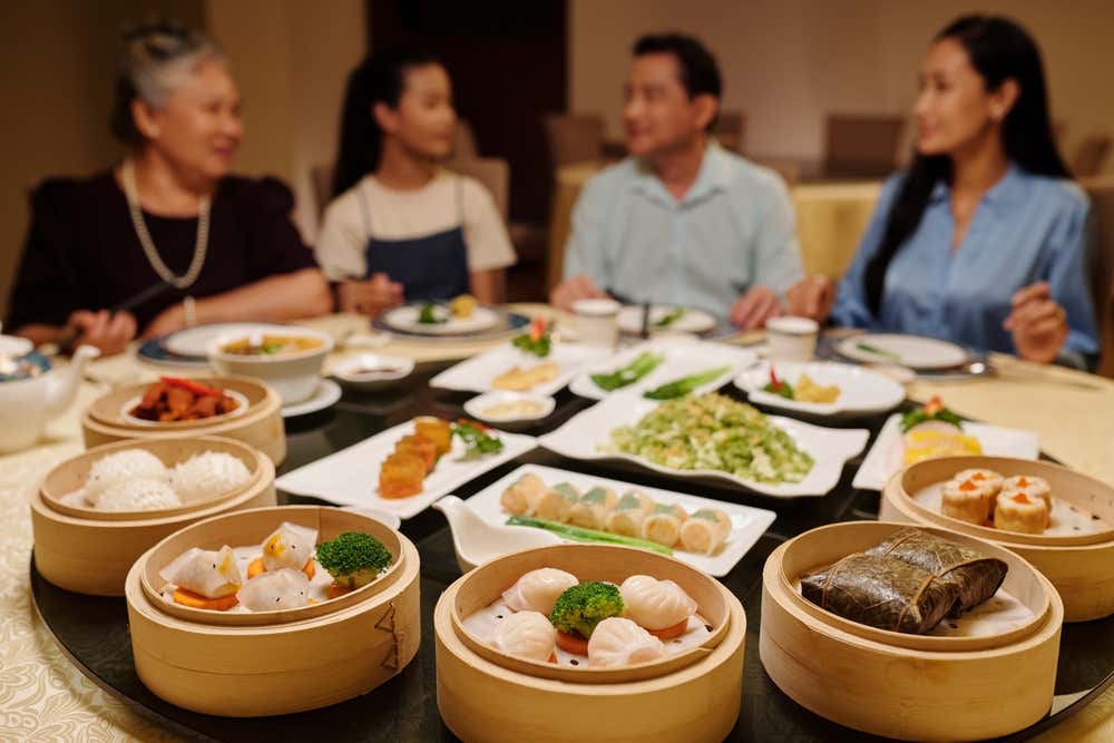 Una famiglia asiatica riunita attorno a un grande tavolo rotondo da pranzo imbandito con numerosi cestelli di bambù e piatti da portata ricchi di dim sum, baozi e pietanze tradizionali