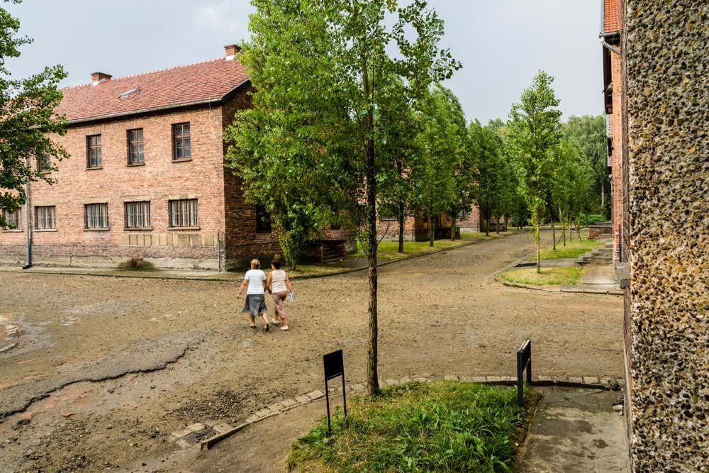 Due visitatrici che camminano lungo le strade sterrate e alberate del memoriale di Auschwitz I, affiancate dagli storici blocchi in mattoni rossi del campo di concentramento.