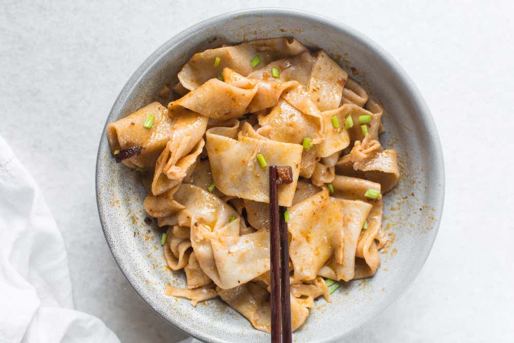 Vista dall'alto di una ciotola grigia contenente noodles cinesi larghi e piatti (biang biang noodles) conditi con salsa piccante all'olio d'aglio e erba cipollina tritata, con un paio di bacchette di legno scuro che ne sollevano uno al centro