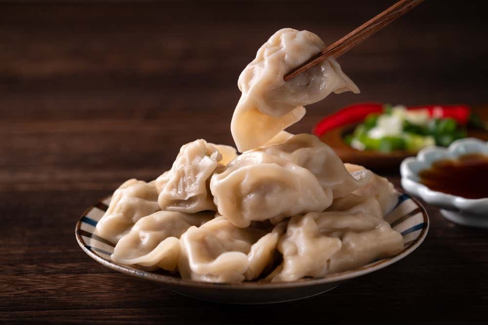 Alt Text (Testo Alternativo)

Alt text: Primo piano di un piatto ceramico colmo di ravioli cinesi al vapore (jiaozi), con bacchette di legno che ne sollevano uno in alto e ciotoline di salsa di soia e scalogno sullo sfondo sfocato