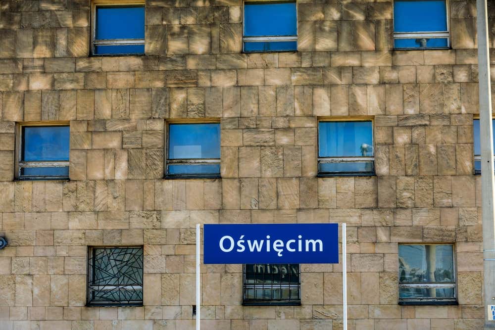 Cartello stradale o ferroviario blu con la scritta bianca "Oświęcim", posizionato davanti alla facciata in pietra con finestre dell'edificio della stazione della città polacca di Oświęcim, nei pressi di Auschwitz.