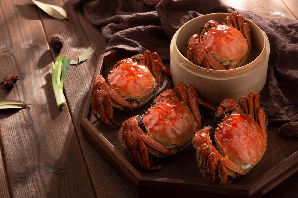 Granchi pelosi cinesi al vapore (hairy crabs) dal carapace arancione brillante disposti su un vassoio di legno e in un cestello di bambù, circondati da aromi come cipollotto e anice stellato su un piano rustico