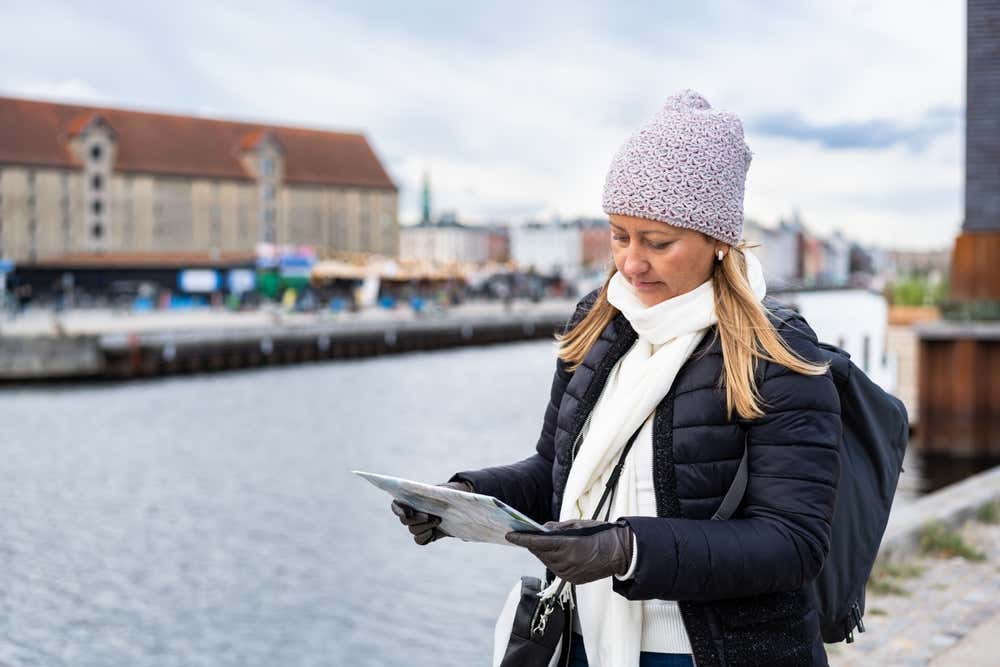 Una donna con berretto di lana, guanti e vestiti invernali studia la mappa di Copenaghen sulle rive di un canale in una giornata nuvolosa.
