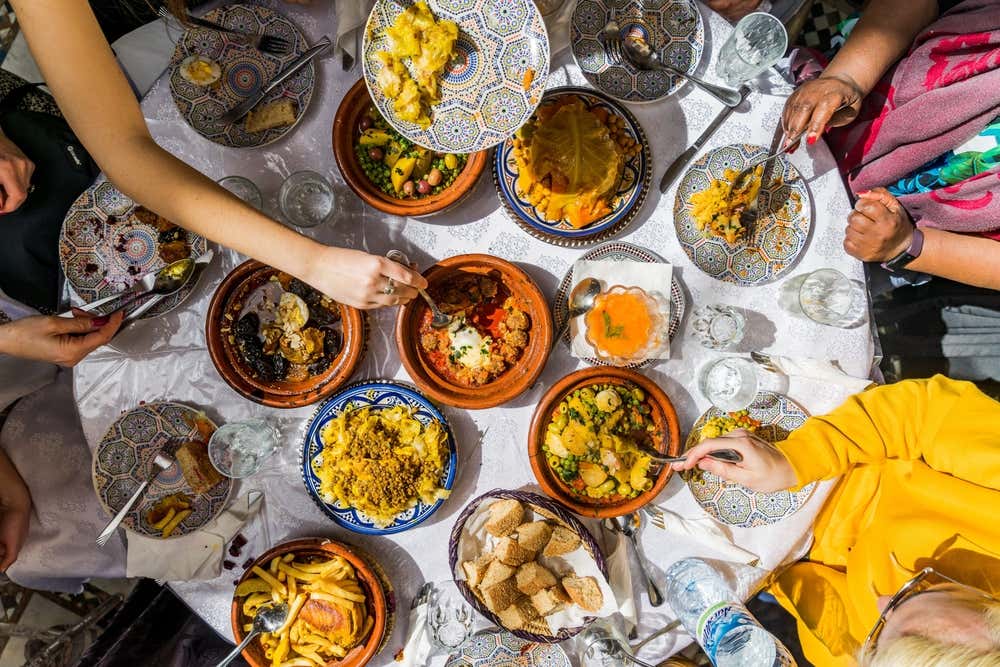 Vista dall'alto di una tavola imbandita con una tovaglia bianca e numerose ciotole piene di pietanze tipiche marocchine dal colore giallo arancione tipico di spezie come la curcuma. Tutto intorno al tavolo sono sedute delle persone di cui si vedono solo le braccia mentre si servono del cibo