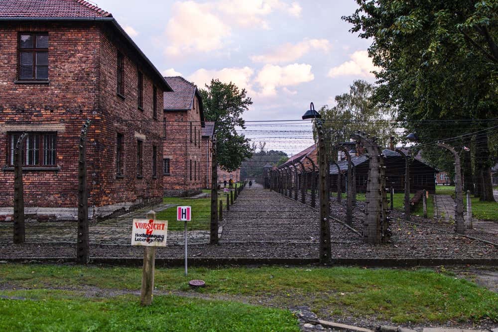 iale interno del campo di concentramento di Auschwitz-Birkenau affiancato da blocchi in mattoni rossi e doppie recinzioni di filo spinato con pali in cemento, con un cartello d'epoca di pericolo in primo piano che cita "Vorsicht